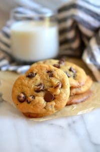 Stack of chocolate chip cookies on parchment paper with glass of milk in background.