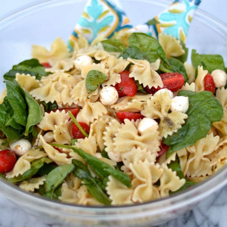 Spinach, tomato and fresh mozzarella pasta salad in a glass bowl with serving spoons.