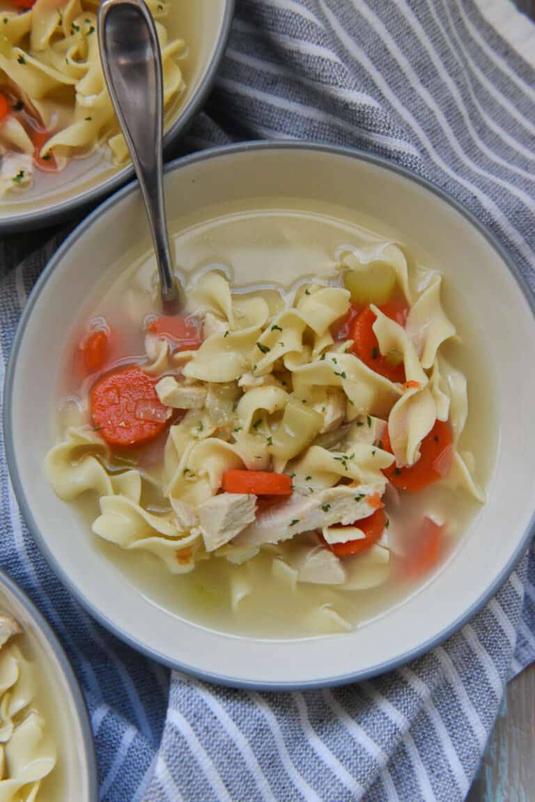 Chicken noodle soup with egg noodles, sliced carrots, celery and onions, garnished with parsley in a gray bowl.