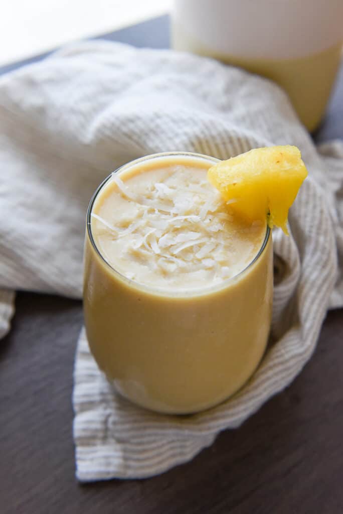 Pina colada protein shake in a glass and garnished with a pineapple chunk and coconut flakes.