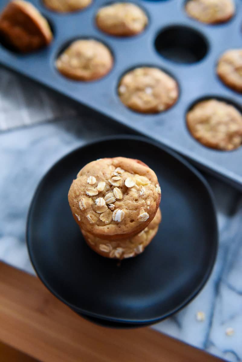 Two Apple Cinnamon Oatmeal Muffins on a black plate stacked on top of one another with muffins in a muffin pan in the background.