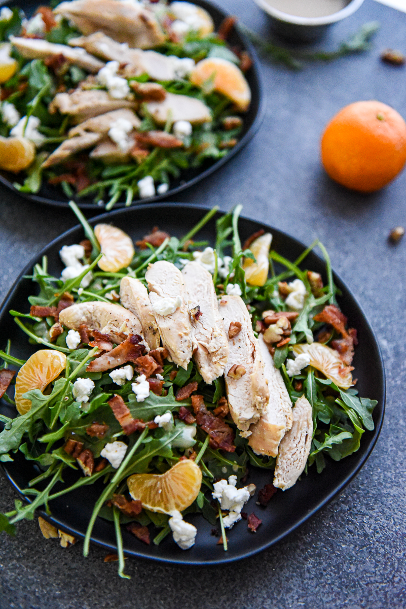 Two arugula salads on black plates with a mandarin orange in the background.