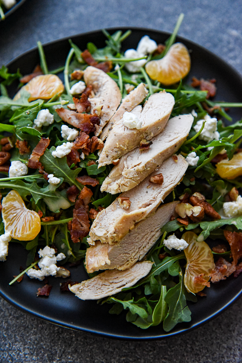 An arugula salad on a black plate topped with bacon, chicken, goat cheese, chopped pecans, mandarin orange wedges and a balsamic dressing.